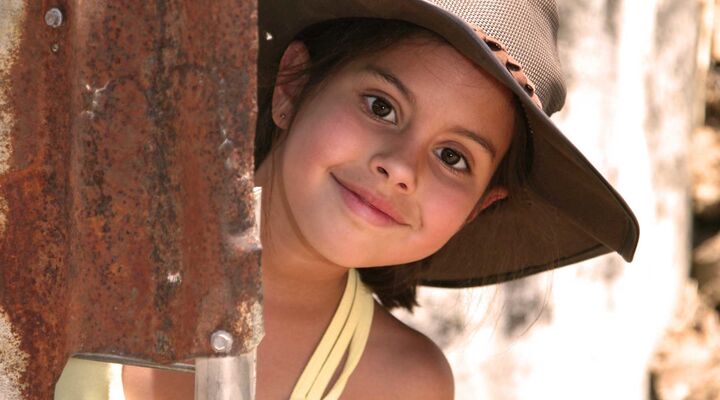 Girl With Dark Eyes And Wide Brimmed Hat Leaning Out Of Rusty Corrugated Iron Wall