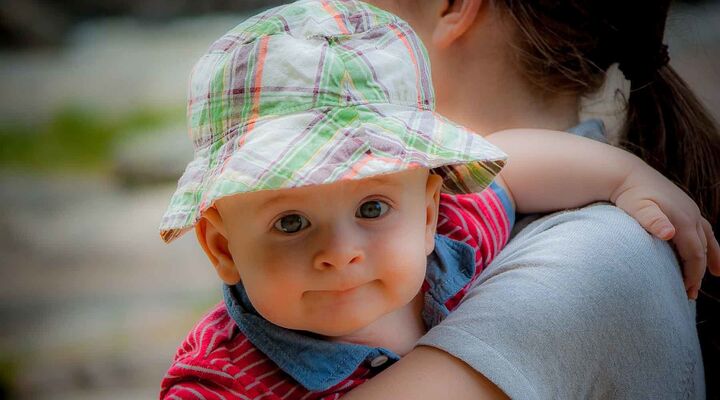 Baby In Red Polo Top And Tartan Hat Looking Over Womans Shoulder